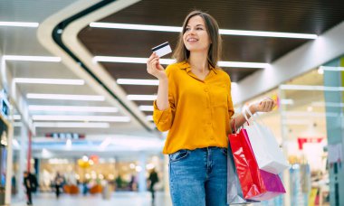 Heyecanlı gülümseyen genç bayan alışveriş merkezinde kredi kartı ve alışveriş torbalarıyla poz veriyor.