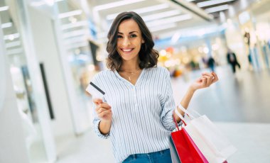 Heyecanlı gülümseyen genç bayan alışveriş merkezinde kredi kartı ve alışveriş torbalarıyla poz veriyor.