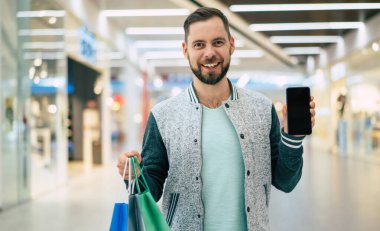 Yakışıklı, genç, şık, sakallı adam siyah cuma günü alışveriş merkezinde yürürken akıllı telefon kullanıyor.