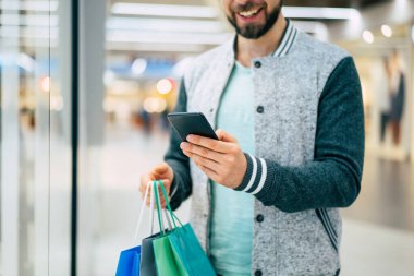 Yakışıklı, genç, şık, sakallı adam siyah cuma günü alışveriş merkezinde yürürken akıllı telefon kullanıyor.
