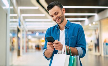 Yakışıklı, genç, şık, sakallı adam siyah cuma günü alışveriş merkezinde yürürken akıllı telefon kullanıyor.