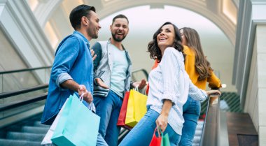 Dört şık gülümseme, alışveriş merkezindeki yürüyen merdivende dikilirken kağıt torbalarla gündelik giysiler içinde heyecanlı modern arkadaşlar eğleniyorlar..