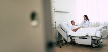 Steteskoplu mutlu, kendine güvenen kadın doktor hastane koğuşundaki hasta adamla konuşuyor.