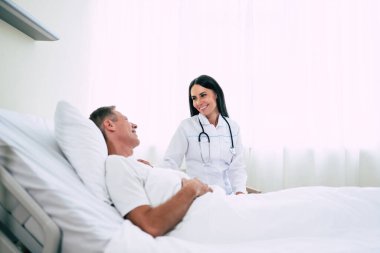 Steteskoplu mutlu, kendine güvenen kadın doktor hastane koğuşundaki hasta adamla konuşuyor.