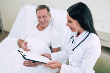 Yakışıklı, olgun, hasta bir adam, klinik yatağında uzanıyor ve beyaz önlüklü, kendine güvenen genç bir doktorla konuşuyor.