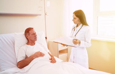 Yakışıklı, olgun, hasta bir adam, klinik yatağında uzanıyor ve beyaz önlüklü, kendine güvenen genç bir doktorla konuşuyor.