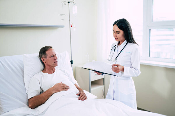 Handsome mature patient man is lying on the clinic bed and has a conversation with a confident young woman doctor in a white coat