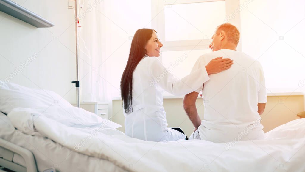 Back view photo of happy doctor and patient while they sitting on the bed in the clinic ward on the window background