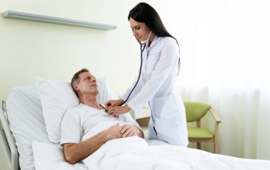 Steteskoplu beyaz önlüklü genç, kendine güvenen kadın doktor hastane koğuşundaki yatakta yatarken hastanın duygularını inceliyor..