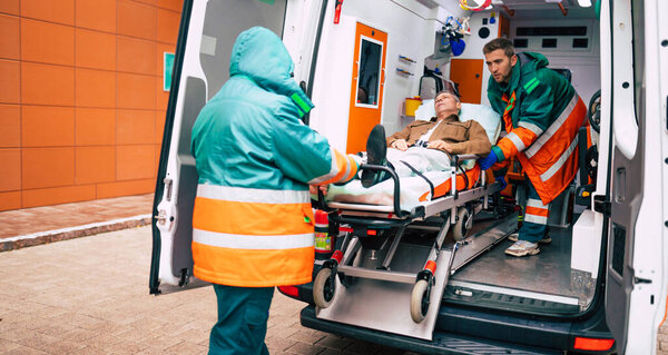 Уверенные и профессиональные врачи выгружают больного пациента из машины скорой помощи на носилках возле здания больницы