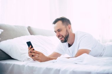 Modern mutlu genç sakallı adam evde yatakta dinleniyor ve telefonda konuşuyor.