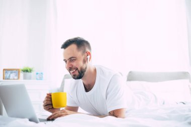 Yakışıklı, modern, sakallı adam sabahları evdeki büyük beyaz yatakta yatarken dizüstü bilgisayarda çalışıyor.