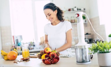 Şirin, güzel ve mutlu esmer kadın mutfakta sebze salatası veya sağlıklı bir içecek hazırlıyor ve eğleniyor.