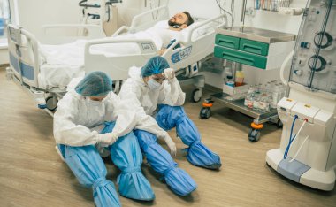 Korunma giysileri içindeki iki bitkin ve çaresiz doktor yerde yatıyor. Koronavirüsü olan bir hastanın arka planında yorgunluk ve hata belirtileri var..
