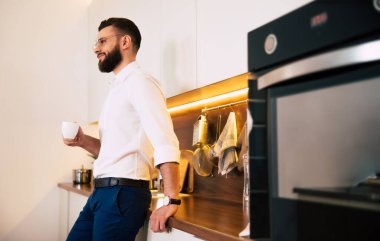 Rahat sakallı, kendine güveni tam bir iş adamı. Sıradan giyiniyor, içki içiyor, kahve kokuyor ve ev mutfağında düşünceli.