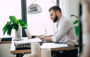 Genç, yakışıklı, sakallı, gözlüklü ve beyaz gömlekli bir iş adamı, bilgisayarında evraklar ve çalışma masasının üstünde kağıtlarla çalışıyor..
