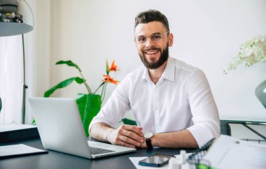 Genç, yakışıklı, gözlüklü, sakallı ve beyaz gömlekli bir iş adamı laptopta çalışıyor. Belgeler masanın üstünde ve kameraya bakıyor..