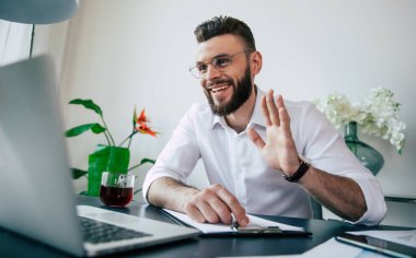 Gözlüklü ve beyaz gömlekli yakışıklı iş adamı dizüstü bilgisayarda çalışıyor ve ekibiyle internette sohbet ediyor..
