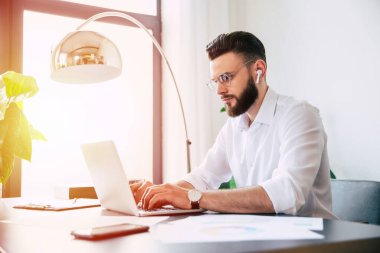 Genç, yakışıklı, sakallı, gözlüklü ve beyaz gömlekli bir iş adamı, bilgisayarında evraklar ve çalışma masasının üstünde kağıtlarla çalışıyor..