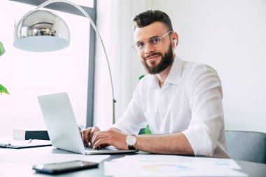 Genç, yakışıklı, gözlüklü, sakallı ve beyaz gömlekli bir iş adamı laptopta çalışıyor. Belgeler masanın üstünde ve kameraya bakıyor..