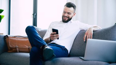 Genç, sakallı, gözlüklü ve resmi giysili bir iş adamı koltukta oturmuş, dinleniyor ve akıllı telefon kullanıyor.