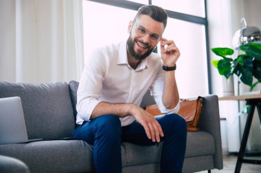 Kendine güvenen, şık, sakallı, gözlüklü ve resmi olarak şık giyinen iş adamı modern ofisteki ya da odadaki kanepede oturuyor.