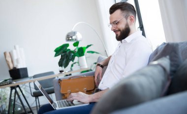 Laptopta çalışırken ve kanepede otururken konsantre ve yakışıklı sakallı bir iş adamının yan görüntüsü.