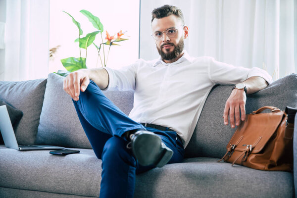 Уверенный в себе стильный красивый бородатый бизнесмен в очках и формальной умной одежде сидит на диване в современном офисе или комнате
