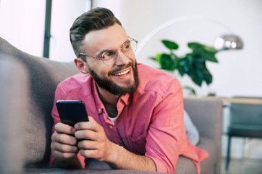 Genç ve mutlu sakallı adam internette sörf yapmak, daktilo etmek, sohbet etmek, oyun oynamak ya da evdeki kanepede dinlenirken bazı uygulamalarla çalışmak için akıllı telefon kullanıyor.