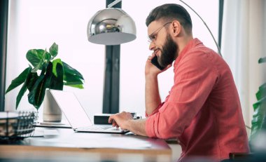 Gözlüklü, kendine güvenen, yakışıklı, sakallı ve şık giyimli adam modern iş yerinde ya da ev ofisinde dizüstü bilgisayarda çalışıyor.