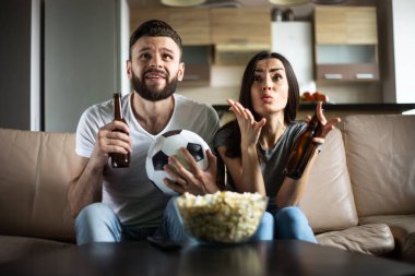 Mutlu taraftar çiftimiz TV 'de futbol maçı izliyor. Yanında abur cubur, bira ve top var.