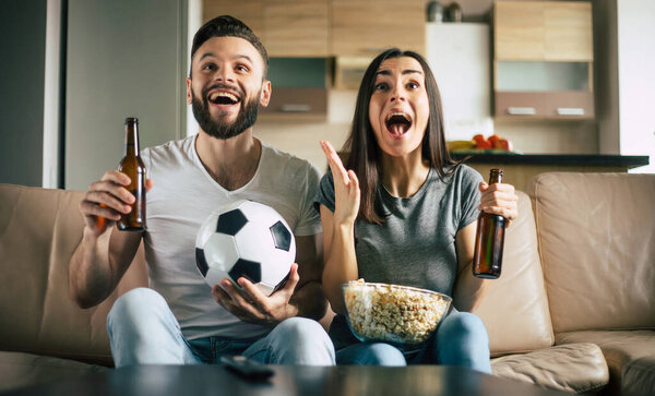 Happy fans couple is watching some football match in TV with snacks, beers and ball on the couch