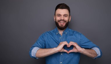 Kot gömlekli, gülümseyen sakallı genç adam kalp şeklinde eller ve kameraya bakıyor.