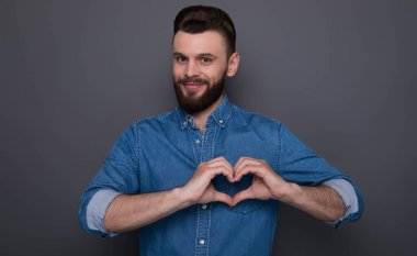 Kot gömlekli, gülümseyen sakallı genç adam kalp şeklinde eller ve kameraya bakıyor.
