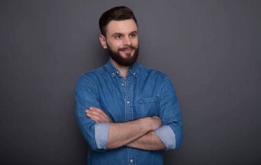 Kollarını kavuşturmuş kot pantolonlu mutlu sakallı adam gri arka planda poz veriyor.