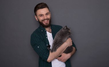 Güzel gülümseyen genç sakallı adam sevimli tüylü kedisini elinde tutuyor ve kamerada en iyi arkadaşlar gibi poz veriyorlar.