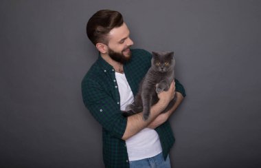 Güzel gülümseyen genç sakallı adam sevimli tüylü kedisini elinde tutuyor ve kamerada en iyi arkadaşlar gibi poz veriyorlar.