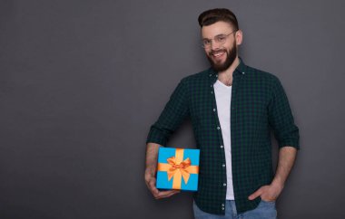 Mutlu, genç, yakışıklı, sakallı bir adam. Keyfi yerinde. Elinde büyük bir hediye kutusu, gri arka planda poz veriyor.