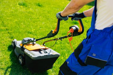 Genç adam arka bahçedeki büyük bir kır evinin yanında özel işçi kıyafetiyle elektrikli çim biçme makinesi kullanarak çimleri biçiyor.