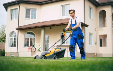 Genç adam arka bahçedeki büyük bir kır evinin yanında özel işçi kıyafetiyle elektrikli çim biçme makinesi kullanarak çimleri biçiyor.