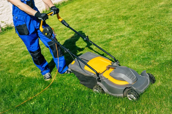 Genç adam arka bahçedeki büyük bir kır evinin yanında özel işçi kıyafetiyle elektrikli çim biçme makinesi kullanarak çimleri biçiyor.