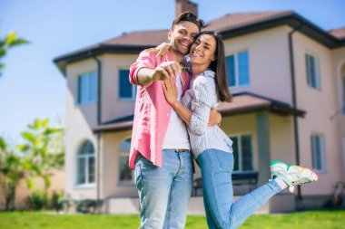Modern, güzel, heyecanlı, birbirine aşık genç bir çift yeni büyük evin önünde ellerinde anahtarlarla durup bu alışverişi kutlarken sarılıyorlar.