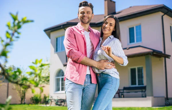 Modern, güzel, heyecanlı, birbirine aşık genç bir çift yeni büyük evin önünde ellerinde anahtarlarla durup bu alışverişi kutlarken sarılıyorlar.