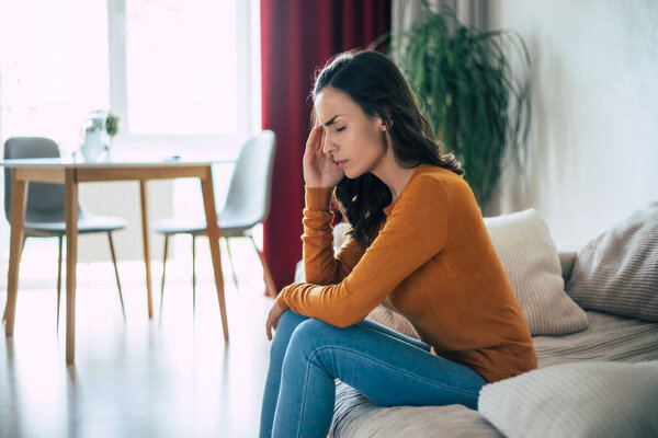  Young sad Brunette woman with severe headache holds her head while she sitting on the couch at home