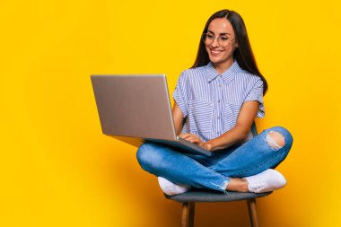 Genç, güzel, modern bir kadın elinde dizüstü bilgisayarla sandalyede otururken sarı arka planda izole bir şekilde oturuyor.