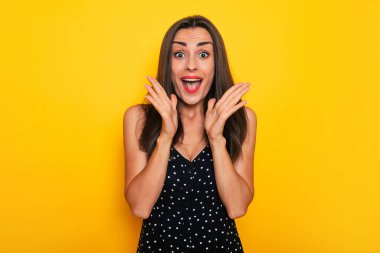 Mutlu, heyecanlı, güzel, gülümseyen, siyah yaz elbisesi giyen esmer kadın sarı arka planda yalnız poz veriyor ve eğleniyor.