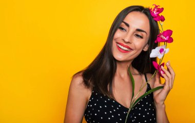 Mutlu, heyecanlı, güzel, gülümseyen, siyah yaz elbisesi giyen esmer kadın sarı arka planda yalnız poz veriyor ve eğleniyor.