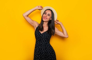 Şapkalı ve elbiseli mutlu genç bayan sarı arka planda poz veriyor ve eğleniyor.