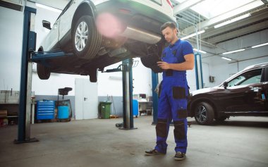 Kendinden emin, genç ve deneyimli araba tamiri uzmanı elinde tabletle arabayı kontrol ediyor ve arıza teşhisi koyuyor.