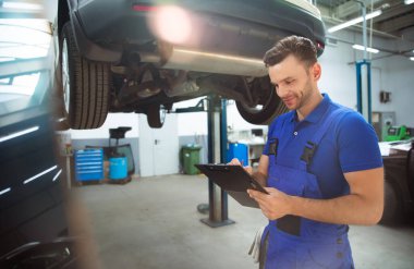 Kendinden emin, genç ve deneyimli araba tamiri uzmanı elinde tabletle arabayı kontrol ediyor ve arıza teşhisi koyuyor.